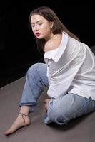 un' bellissimo ragazza nel un' bianca camicia e jeans si siede su un' grigio sfondo. foto