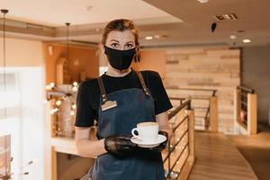 una cameriera che indossa una maschera nera e guanti usa e getta serve i clienti in un bar foto