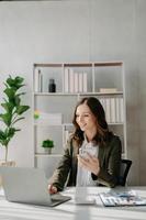 fiducioso attività commerciale esperto attraente sorridente giovane donna digitando il computer portatile ang Tenere digitale tavoletta su scrivania nel creativo casa ufficio. foto
