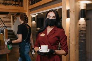 una donna manager di un ristorante indossa una maschera nera e guanti usa e getta in possesso di una tazza di caffè in un ristorante foto