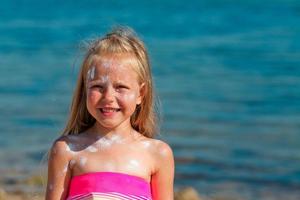 un' bellissimo poco ragazza sorrisi nel un' costume da bagno in piedi di il mare con protezione solare spalmato su sua viso. foto