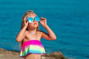 bellissimo poco ragazza nel un' costume da bagno e occhiali da sole sta di il mare con protezione solare spalmato su sua corpo. foto