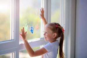 bambini ragazza disegnare con palme su il finestra. dipinto mani partire un' marchio su il bicchiere. quarantena. restare casa. foto