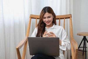 ritratto di un' bellissimo giovane donna nel un' bianca camicia Tenere un' bicchiere di latte e un' il computer portatile per genere sua proprio storia e seduta su un' di legno sedia dentro il Casa, il concetto di riposo foto