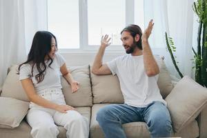 un' uomo e un' donna seduta a casa su il divano nel bianca elegante magliette e chat allegramente sorridente e ridendo a casa. maschio e femmina amicizia foto