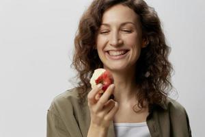 contento sorridente Riccio bellissimo donna nel casuale cachi verde camicia mangiare Mela con un appetito considera esso in posa isolato su al di sopra di bianca sfondo. naturale eco-friendly prodotti concetto. copia spazio foto