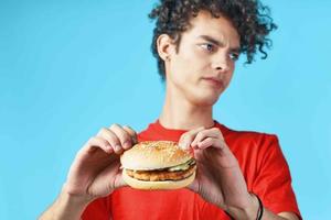 allegro tipo con Riccio capelli nel un' rosso maglietta con un' Hamburger nel il suo mani veloce cibo dieta foto