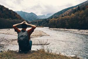 donna escursionista si siede su il fiume banca e ammira il montagna paesaggio nel il distanza indietro Visualizza foto