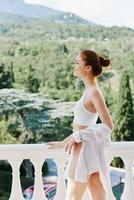 ritratto di giovane bellissimo donna nel un' bianca camicia ammira il verde natura su il balcone soleggiato giorno foto