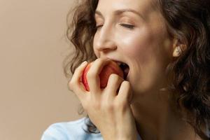 carino contento Riccio bellissimo femmina nel jeans casuale camicia morsi Mela godere dolce frutta in posa isolato su al di sopra di beige pastello sfondo. salutare cibo. naturale eco-friendly prodotti concetto. copia spazio foto