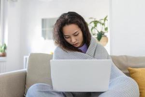 asiatico donna avvolto nel un' accogliente coperta seduta su il divano e Lavorando su sua il computer portatile foto