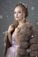 bellissimo modello con alla moda trucco nel un' pelliccia cappotto. lusso inverno ragazza su un' grigio sfondo disseminato con neve. foto