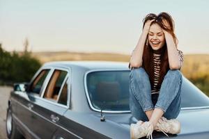 un' giovane donna si siede su il cappuccio di sua auto e ride con sua occhi Chiuso, un' viaggio a partire dal un' rumoroso città per natura nel estate, viaggio stile di vita foto
