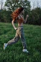 un' donna con lungo rosso capelli lavori su e corre su il verde erba nel il parco nel pantaloni della tuta e scarpe da ginnastica foto
