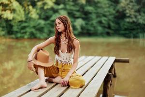 un' giovane hippie donna si siede su un' lago ponte indossare elegante eco Abiti e sorridente foto