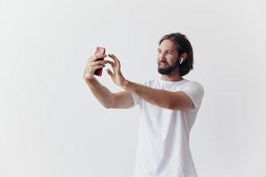 un' maschio blogger videoregistrazione lui stesso su il suo Telefono e chat con persone in linea con un' Sorridi nel un' bianca maglietta contro un' bianca parete foto