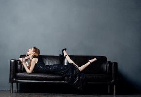 attraente donna nel nero vestito bugie su un' pelle divano in casa con sollevato gambe foto