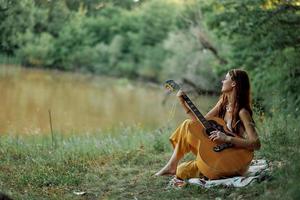 un' hippie donna giocando sua chitarra sorrisi e canta canzoni nel natura seduta su un' plaid nel il sera nel il tramonto luce del sole. un' stile di vita nel armonia con il corpo e natura foto