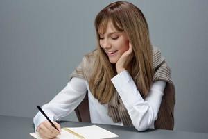 contento giovane bionda alunno giornalista signora nel caldo maglione scrittura amore lettera articolo saggio seduta a posto di lavoro nel grigio moderno casa ufficio. formazione scolastica e carriera professione concetto. copia spazio foto