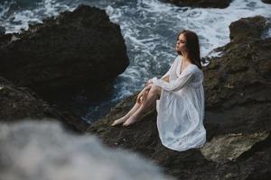 un' donna nel un' bianca nozze vestito è seduta a piedi nudi su un' onda scogliera inalterato foto