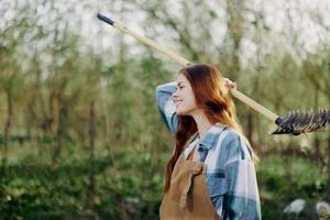 un' donna sorridente meravigliosamente e guardare a il telecamera, un' contadino nel opera Abiti e un grembiule Lavorando all'aperto nel natura e Tenere un' rastrello per raccogliere erba e foraggio per il animali nel il giardino foto