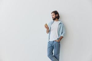 libero professionista del Millennio uomo con un' barba potabile caffè a partire dal un' riciclato tazza nel elegante fricchettone Abiti bianca maglietta blu jeans e camicia su un' bianca sfondo foto