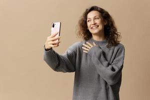 amichevole Riccio bellissimo femmina nel grigio casuale maglione fare video chiamata con Telefono in posa isolato su al di sopra di beige pastello sfondo. sociale media, Rete, distanza comunicazione concetto. copia spazio foto