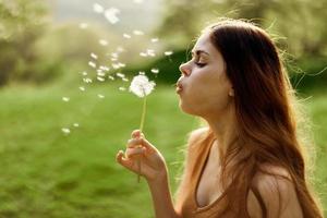 donna con un' dente di leone fiore nel sua mani sorridente e soffiaggio su esso contro un' sfondo di estate verdura e luce del sole foto