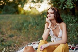 ragazza vestito come un' hippie eco rilassante nel il parco, seduta su un' coperta nel il tramonto, rilassato stile di vita foto