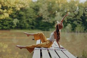 un' hippie donna si siede su un' ponte su il banca di un' fiume e gode il bellissimo scenario in giro suo, contento nel il tramonto autunno sole. ecologico stile di vita foto