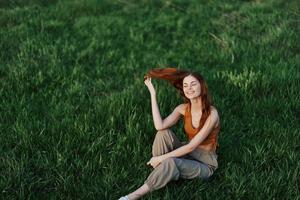 un' donna godendo il all'aperto seduta nel il parco su il verde erba nel casuale capi di abbigliamento con lungo fluente capelli, illuminato di il luminosa estate sole senza zanzare foto