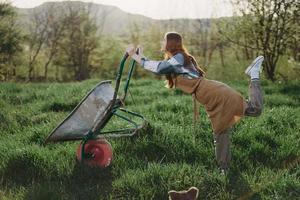 un' giovane donna rotoli un' giardino carrello con suolo per piantare nel sua verde natura giardino e sorrisi foto