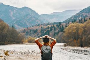 un' viaggiatore nel un' maglione cappello con un' zaino gesti con sua mani e rilassa su il fiume banca nel il montagne foto