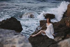 donna nel bianca vestito si siede su un' pietra vicino il oceano onde viaggio natura foto