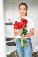 ritratto di giovane donna nel bianca maglietta con mazzo di rosso fiori nel mani vicino finestra nel luminosa cucina a casa foto