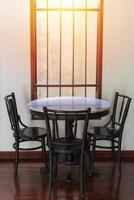tavolo rotondo in legno e sedie vicino a una finestra con raggi di sole su un pavimento in legno e un muro bianco foto