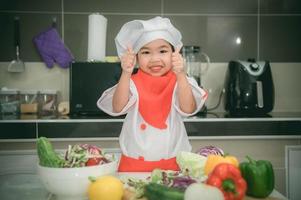 carino asiatico ragazza indossare capocuoco uniforme con un' lotto di vegatable su il tavolo nel il cucina stanza, fare cibo per mangiare cena, divertente tempo per bambini foto