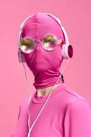 un' pazzo concettuale ritratto di un' giovane donna senza un' viso maschera su un' rosa sfondo e indossare brillante occhiali da sole ascoltando per musica su sua cuffia. il concetto di un' nuovo mondo e cyberspazio foto