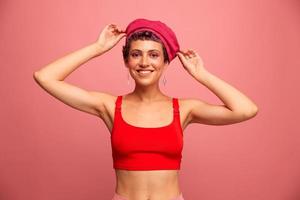 giovane atletico donna con un' corto taglio di capelli e viola capelli nel un' rosso superiore e un' rosa cappello con un atletico figura sorrisi e smorfie mostrando segni con sua mani guardare a il telecamera su un' rosa sfondo foto