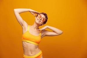 giovane donna nel un' luminosa giallo fitness tuta da ginnastica con un Aperto pancia e occhiali da sole su un arancia sfondo sorridente foto