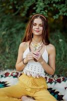 giovane hippie donna seduta nel natura su un' plaid con un' fiore nel sua mani e guardare a il telecamera sorridente Cordiali saluti nel eco-abbigliamento nel autunno nel il tramonto foto