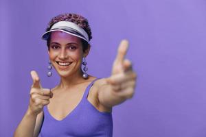 ritratto di un' sportivo moda donna in posa sorridente con denti e puntamento un' dito a il telecamera nel un' viola yoga tuta da ginnastica e un' trasparente berretto su un' viola monocromatico sfondo foto