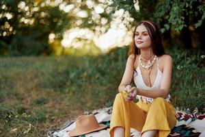 ragazza vestito come un' hippie eco rilassante nel il parco, seduta su un' coperta nel il tramonto, rilassato stile di vita foto