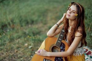 hippie eco amichevole donna con un' chitarra rilassante nel natura seduta su un' plaid di il lago nel il sera nel il raggi di il ambientazione sole. un' stile di vita nel armonia con il corpo e natura foto