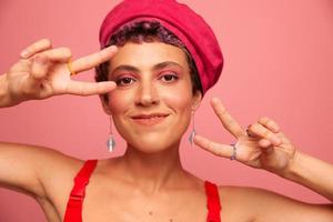 giovane atletico donna con un' corto taglio di capelli e viola capelli nel un' rosso superiore e un' rosa cappello con un atletico figura sorrisi e smorfie mostrando segni con sua mani guardare a il telecamera su un' rosa sfondo foto