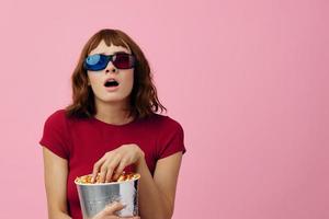 confuso concentrato carino testa Rossa signora nel rosso maglietta 3d bicchieri mangiare Popcorn orologio film in posa isolato su al di sopra di rosa studio sfondo. copia spazio striscione. moda cinema concetto. divertimento offrire foto