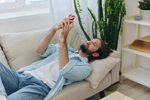 un' uomo con un' barba bugie su il divano durante il giorno a casa e sembra a il suo Telefono rilassante su il suo giorno spento, un' uomo gioco d'azzardo su il azione mercato in linea su il suo Telefono foto