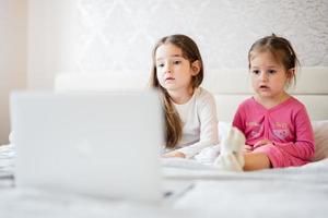 Due carino poco sorelle indossare pigiama guardare cartone animato di il computer portatile nel mattina a casa. foto