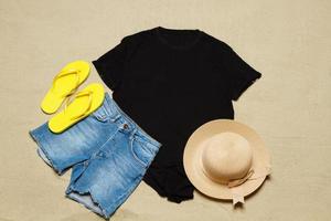 sabbia spiaggia struttura sfondo. modello nero estate maglietta copia spazio. vuoto modello donna camicia superiore Visualizza. estate Accessori cappello, jeans, infradito. piatto-lay avvicinamento maglietta spiaggia. tempo di spiaggia foto