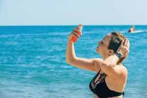 bellissimo giovane bionda donna nel un' costume da bagno rinfresca e idrata sua viso con un antiabbronzante Tonico mentre su il spiaggia sotto il luminosa raggi di il estate sole, copia spazio foto